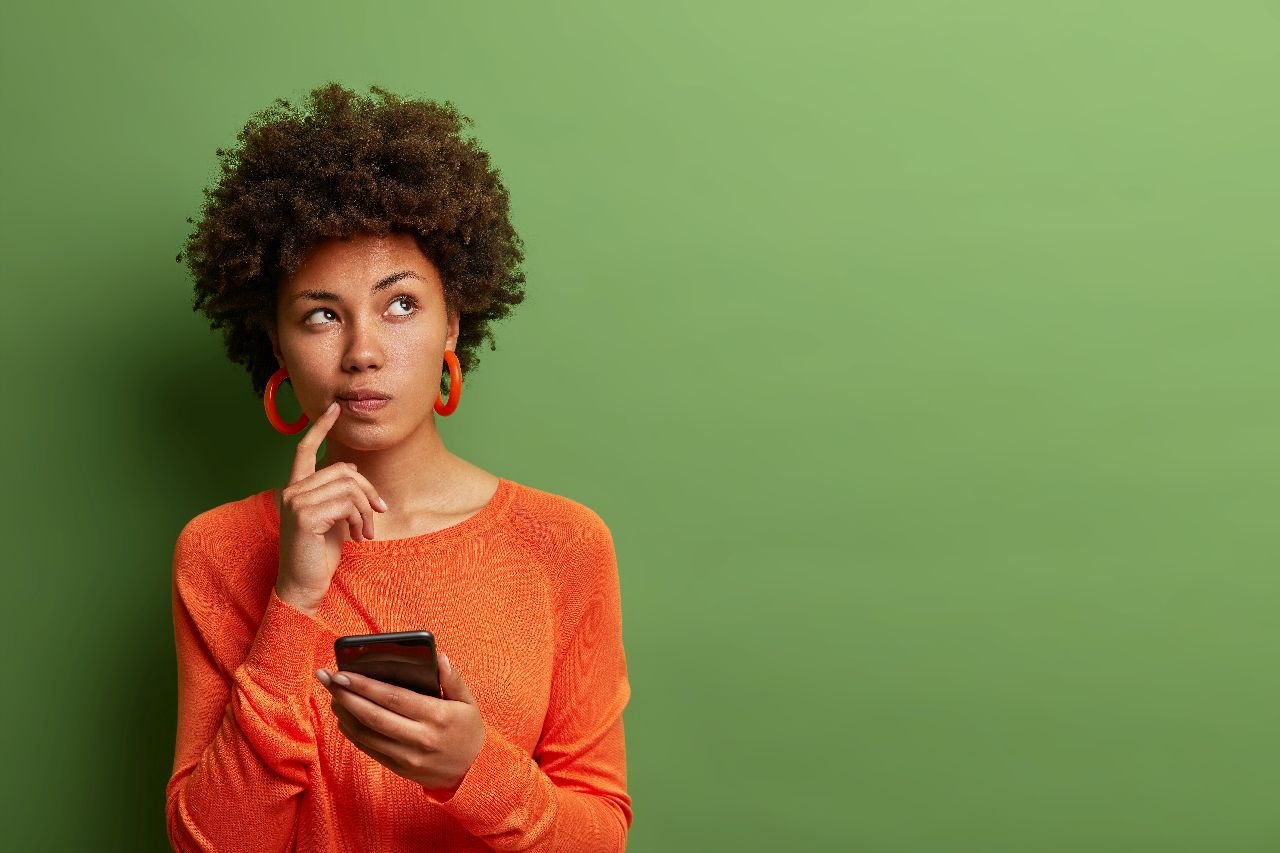 Young woman standing against green background looks curious while holding phone