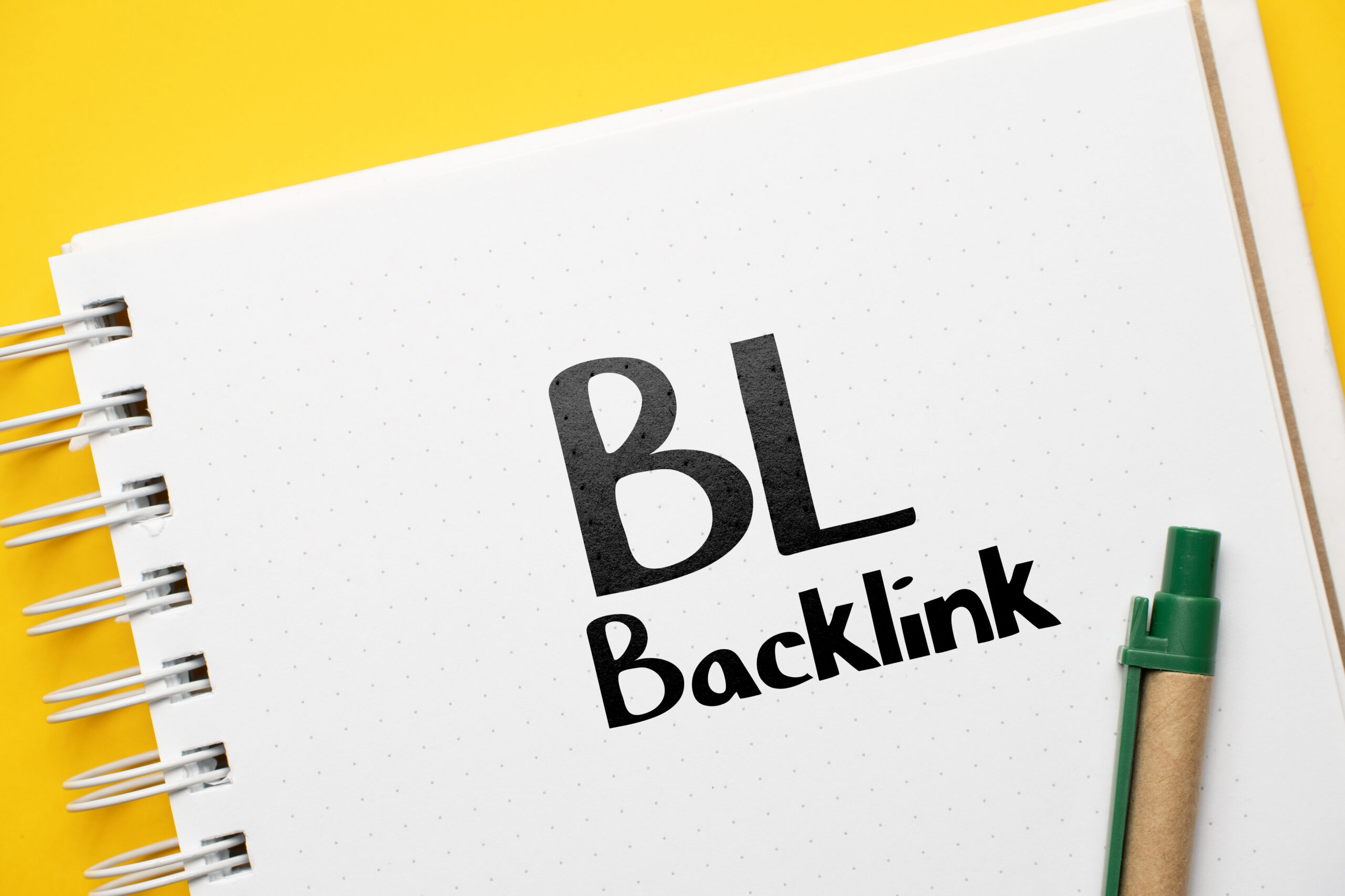 A open note book with a pen next to it. On the open, the letters "BL" are written in marker with the word "backlink" below it.