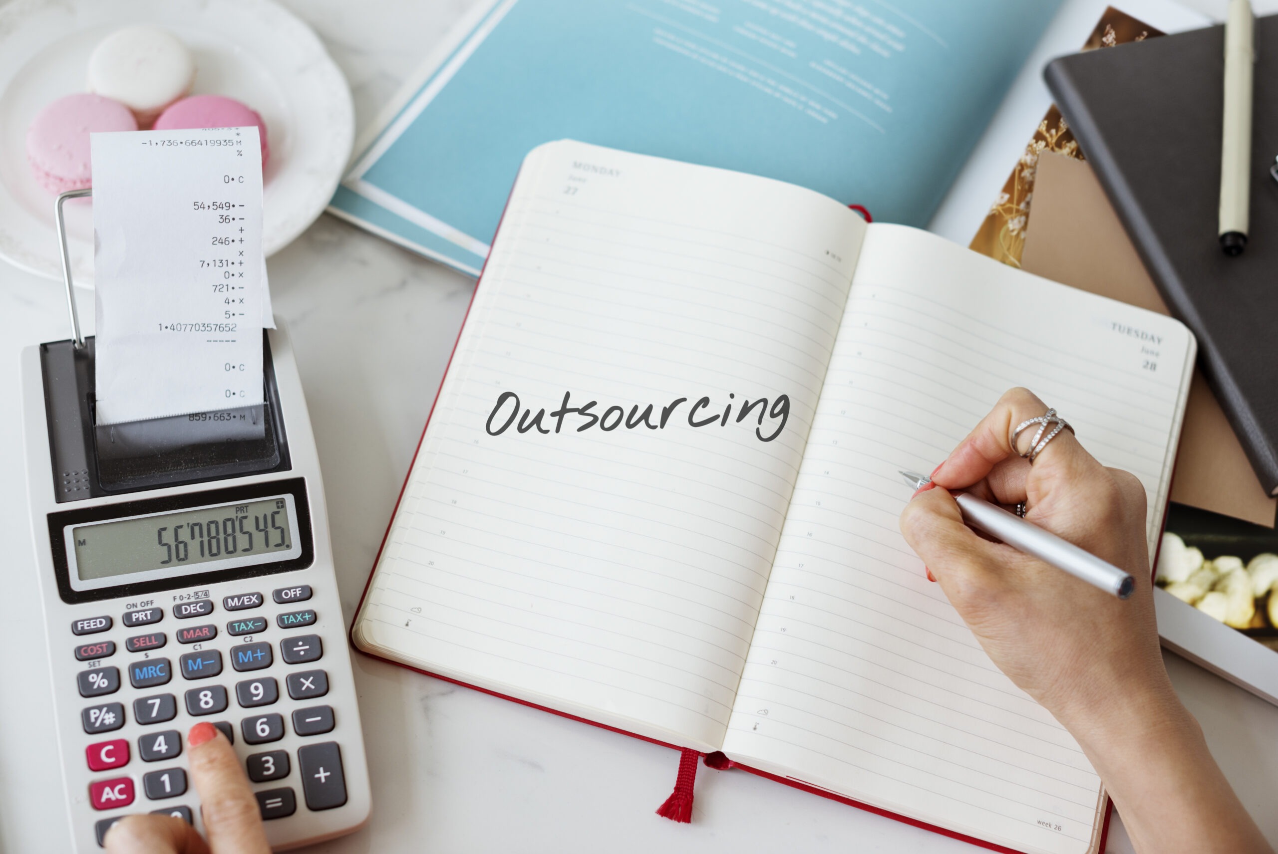 A person writing with a pen on a notebook that says, "outsourcing" on the other paper while using a calculator.