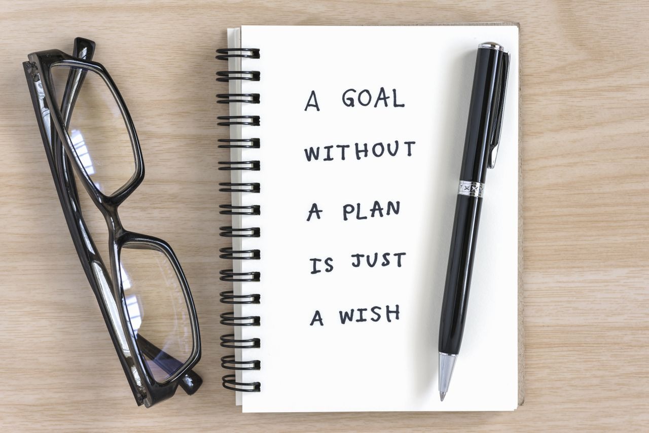 Notebook laying on a table with a pen placed on it. Handwritten words say "A goal without a plan is just a wish."