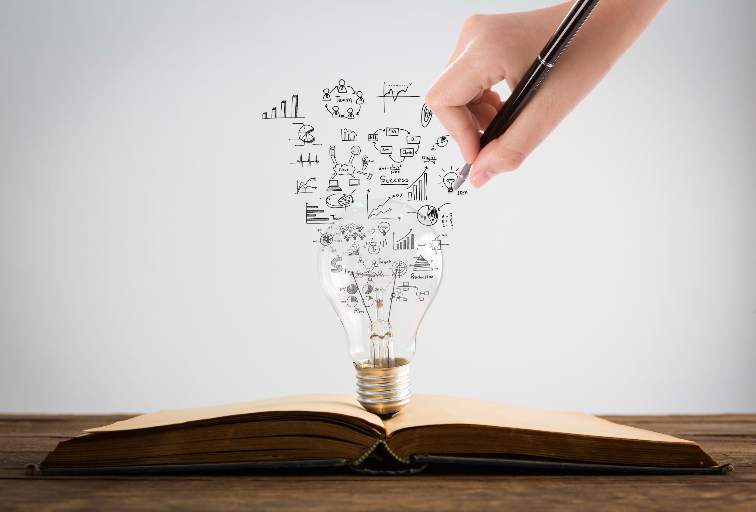 Journal laying open on a table with a lightbulb standing up in the middle. A hand is drawing SEO symbols with a pencil behind the bulb.