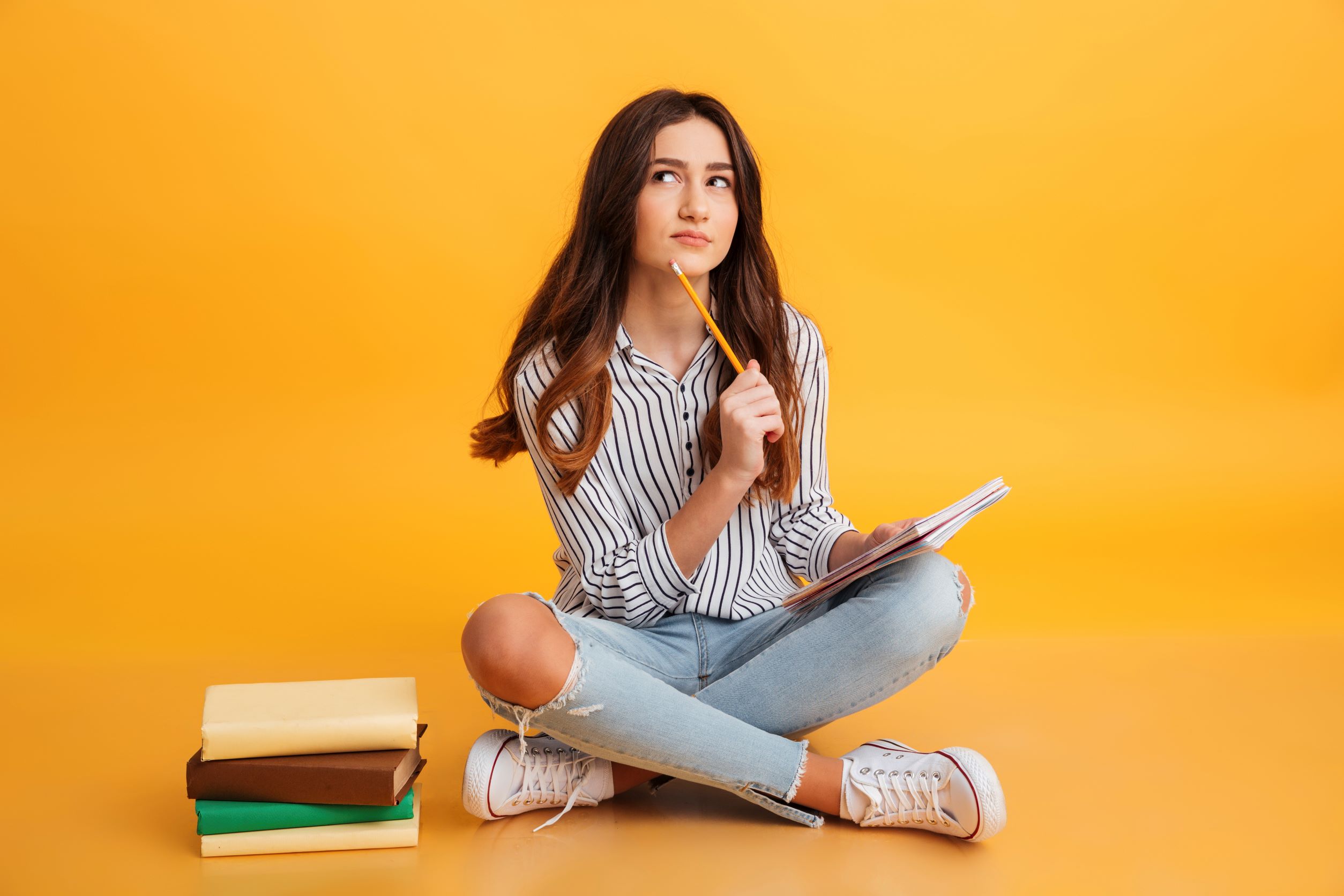 Young woman sitting cross-legged, with a pencil and paper and a stack of books, looking pensive
