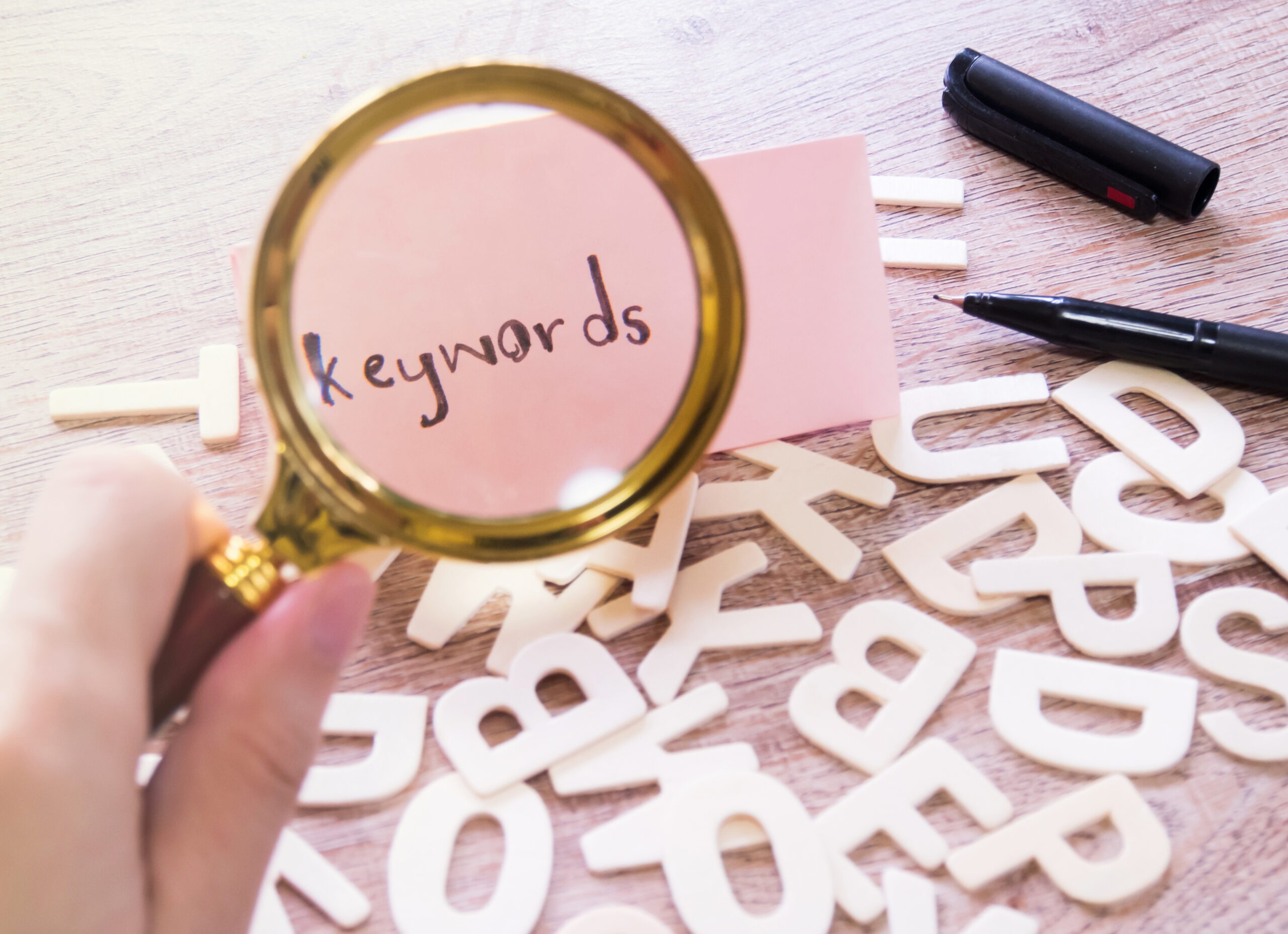 A hand holding a magnifying glass on to a small pink paper that has the word, "keyword" hand written with letters cut out sporadically all over the table. 